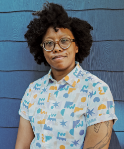 Outdoor photo portrait of a Black woman with dark hair, round glasses, a colorful blouse and tattoos on her arm standing in front of an aqua house exterior