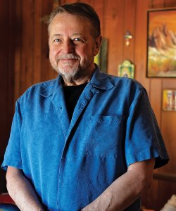 An older man with brown hair and a graying goatee stands in a warm-toned living room with a twinkle in his eye. He wears a bright blue short-sleeved button-up and bracelets.