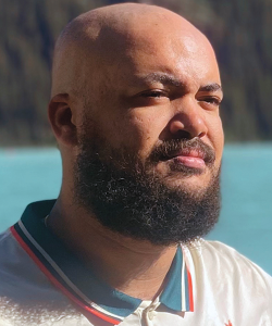 A bald Black man with a beard looks into the distance. He wears a white shirt with a blue-and-red striped collar and a body of water can be seen in the background.