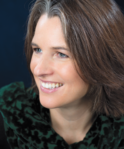 The Author’s Wife Is Also an Author: On the Fantasy and Reality of Writing and Partnership A photo portrait of Erin Almond, a middle-aged white woman with medium length brown hair and blue eyes, smiles while looking slightly off to the side. She wears a dark green top and is posed against a dark background.