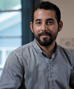 A photo portrait of Mosab Abu Toha, a Palestinian man with olive skin and short dark hair. He wears a gray collared shirt and sits indoors with a window in the background.
