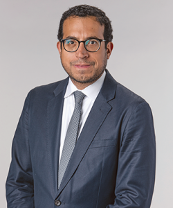 A professional portrait of Gilbert Cruz, a Latino man with tan skin, short brown hair, and black glasses. He wears a dark blue-gray suit with a white shirt and gray patterned tie.
