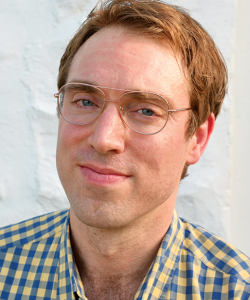 Portrait of a white man with red hair and glasses. He smiles with closed lips and wears a checkered flannel.