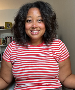Indoor photo portrait of a woman with wavy dark hair and red and white striped T-shirt sitting at a desk