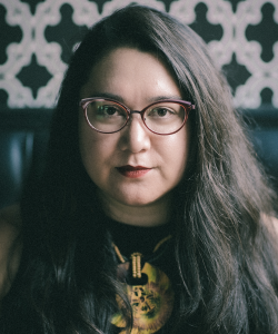 A photo of Janine Joseph, a white woman with long, dark hair and glasses. She wears a black graphic tee with a yellow image and sits in a room with a black and white patterned wallpaper.