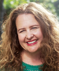 A sunny photo portrait of Jennifer Dickinson, a middle-aged white woman with dark blonde curly hair. She wears a green eyelet top and red lipstick with a smile.