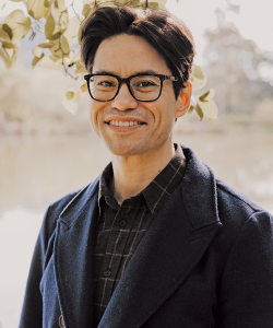 First Fiction 2025 A photo portrait of Jon Hickey, an Indigenous man with dark brown hair, light medium skin, and black square glasses. He smiles and stands in front of a flowering tree. He wears a navy blue peacoat and a dark collared shirt.