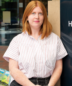 A white lady with red hair and blue eyes stands outside the Hub City Bookshop. She wears a pinstriped shirt and black jeans.