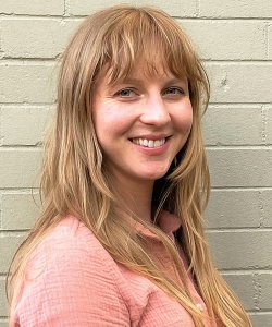 A white woman with long blonde hair and bangs wears a pink shirt against a white brick wall.