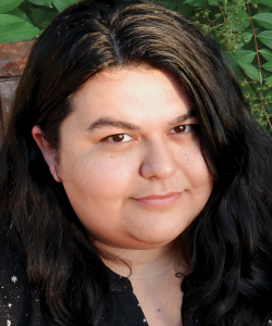 A photo portrait of an Indigenous woman with long black hair and a black star-patterned shirt. She smiles up at the camera and sits against a background of nature.