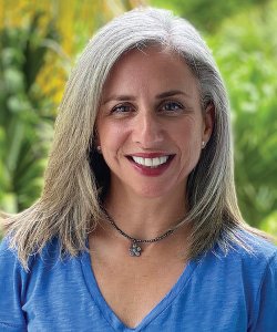 Brenda Ferber, a white woman with tan skin and blonde hair, smiles at the camera against a background of green.