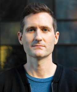 A photo portrait of Michael Deagler, a white man with short brown hair, light skin, and blue eyes. He wears a dark shirt and a shadow falls slightly over the left side of his face.