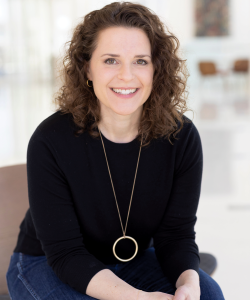 A photo of Michelle Wildgen, a white woman with brown curly hair and brown eyes. She wears a black long-sleeved shirt, a necklace with a round ring pendant, and blue jeans. She sits with her hands folded and smiles brightly.