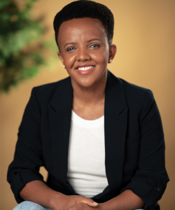 Mihret Sibhat, a Black woman with natural hair, smiles brightly. She wears round stud earrings, a black blazer, distressed jeans, and a white shirt.