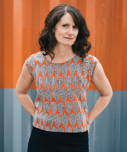 A portrait of Mira Rosenthal, a white woman with light skin and dark brown hair. She wears an orange top with a black and white leaf pattern on top of a black skirt. She poses with hands on hips.