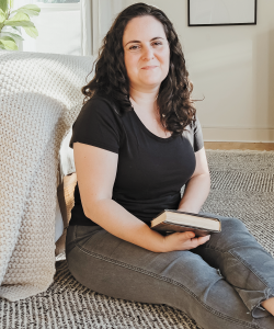 A white woman with wavy dark brown hair sits on the floor next to her bed. She wears a black t-shirt and gray jeans cropped at the ankle. She holds a book in her lap. The woven bedspread is a light cream color and the rug on the floor is black and white.