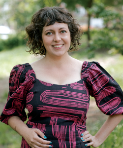 Nicole Haroutunian, a woman with light skin and short wavy brown hair, smiles. She wears a pink and black patterned dress with puffed sleeves and stands in a park.