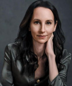 A photo portrait of Nicole Graev Lipson, a white woman with long, dark brown hair. She wears a grey silk blouse and a necklace with a gold pendant. Her left hand rests slightly on her neck.