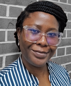 In the Shadow of a Homeplace: One Writer’s Life in Mississippi Olufunke Ogundimu, a Black woman with braids coiled around her head, smiles slightly. She wears clear rimmed glasses, a blue and white striped shirt, and silver earrings. She stands against a gray brick wall.
