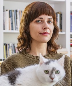 A portrait of Paige Lewis wearing a green-brown sweater, with a white cat with gray markings sitting in the foreground, looking at the viewer.