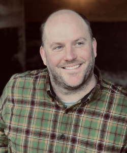 Photo portrait of white man with short, dark facial hair and plaid shirt