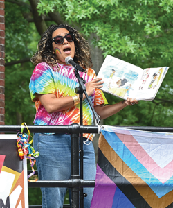 Raakhee Mirchandani, a South Asian woman with big sunglasses and a rainbow tie-dyed shirt, reads from a picture book while standing on a stage in a park. Posters to protest the book ban and an LGBTQIA+ flag are attached to the stage.