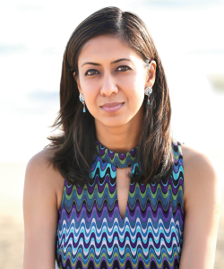 A photo portrait of Namrata Poddar, a South Asian woman wearing pencil eyeliner and dangly silver earrings with a blue patterned dress.