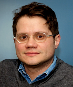 A photo portrait of Ricardo Alberto Maldonado, a Puerto Rican man with brown hair, light medium skin, and a square jaw. He wears round clear-rimmed glasses, a blue collared shirt, and a gray v-neck sweater. He is posed against a blue background.