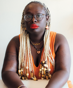 Brittany Rogers, a dark-skinned Black woman wearing thin black-rimmed glasses and orange-red lipstick. She has blonde hair woven into her braids and dangly earrings.