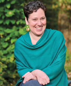 A photo portrait of Sarah Yahm, a white Jewish woman with short curly hair and a big smile. She wears a loose turquoise knit top and gold statement earrings. She sits outdoors with trees in the background.