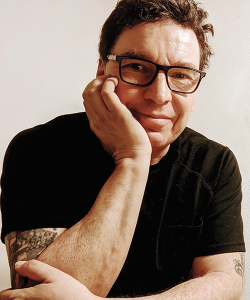 Tommy Archuleta, a man in his late fifties, sits with his face leaning against his right hand. He wears black-rimmed glasses and a black t-shirt and has slightly tousled brown hair. On both arms, the black outlines of tattoos are partially visible.