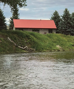 Outdoor photograph of a small house with a red roof sitting atop a river with grassy banks
