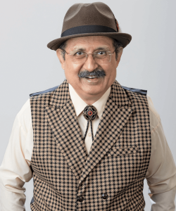 Indoor photo portrait of man with a gray mustache wearing a fedora, rimless glasses, a checked vest, and a bolo tie