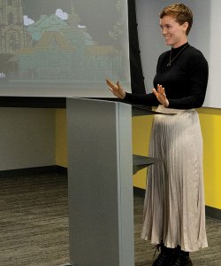 A white woman wearing a black shirt and tan skirt stands in front of a projector screen and whiteboard with her hands lifted outward.