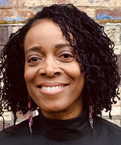 A Black woman with shoulder-length micro locs smiles. She stands against a brick wall and wears beaded earrings and a black turtleneck.