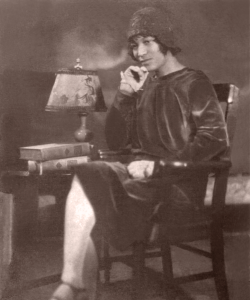An archival photo portrait of Zora Neale Hurston, a Black woman sitting in a chair wearing a bucket hat and a dark velvet dress. She has a pensive look on her face with one hand on her chin.