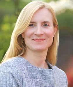 A woman with long blonde hair, wearing a textured jacket, smiles at the camera. Sarah McGrath