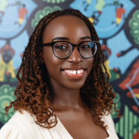 Headshot of Kukuwa Fraswer, wearing glasses and a white shirt, who smiles in front of a wall with a brightly painted mural