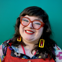 Headshot of Paloma Hernando, who smiles in a colorful outfit with bright red glasses, standing before a turquoise wall