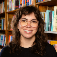 Kate McMullen, a person with brown curly hair and glasses, smiles as she stands in front of a bookshelf