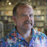 Headshot of Ander Monson Headshot of Ander Monson, a man with a mustache and a Hawaiian shirt who stands before some bookcases