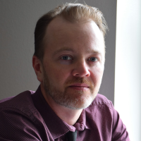 Headshot of Jeremy Schraffenberger, who wears a maroon colored shirt and sits with half his face in shadow and half in sunlight