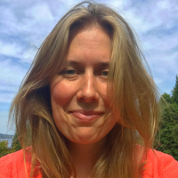 Head shot of Naomi Gibbs, a person with blond hair hanging on either side of her face, wearing a red shirt as she stands before a blue sky and trees
