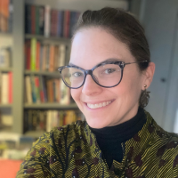 Head shot of Suzanna Tamminen, a person with brown hair and glasses who is smiling in front of a wall of books