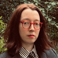 Head shot of Camille Ralphs, wearing glasses and a serious expression while sitting in a walled garden