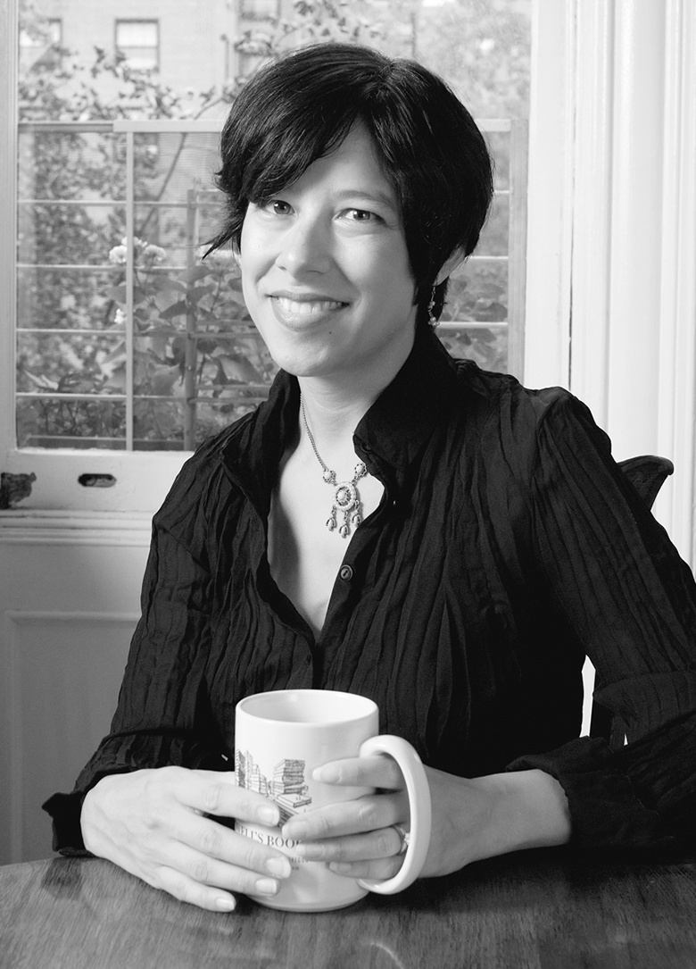 black-and-white portrait of a woman with dark hair and dark-color blouse and metallic necklace sitting in front of a window and holding a mug in her hands