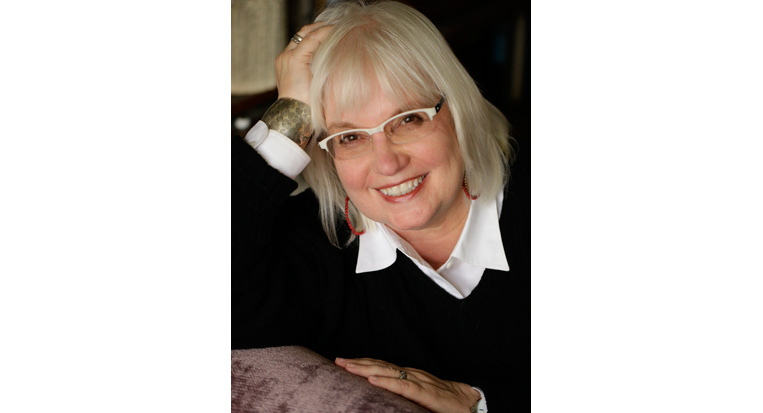 Susan Larson with white-framed glasses and light shoulder length hair smiling and leaning on her arm wearing a black sweater over a white collared shirt.