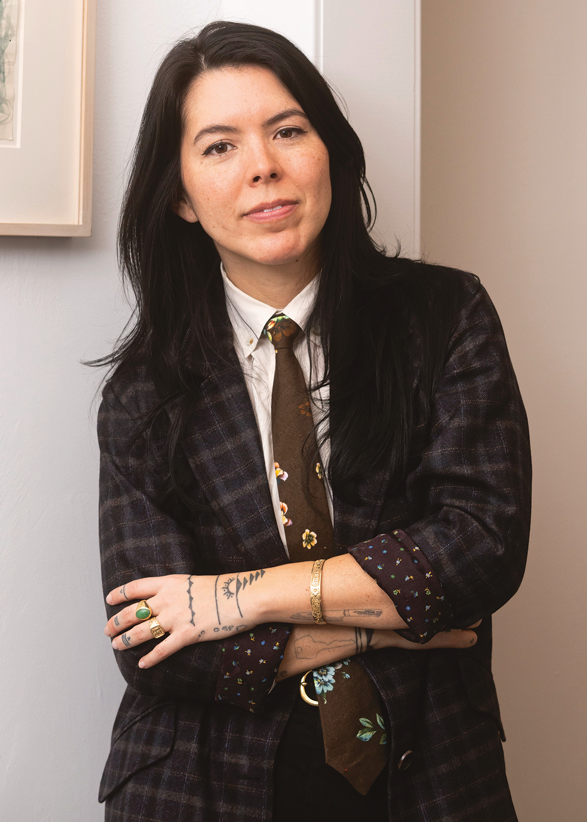 Indoor photo portrait of a woman with long dark hair and tattoos on her arms wearing a dark plaid jacket and tie