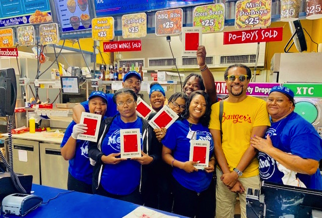 Colson Whitehead in a bright yellow t-shirt with staff at Melba's holding his book and smiling.